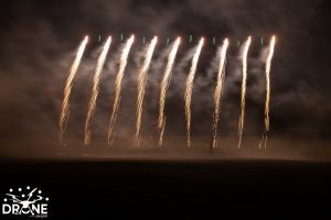 Vertical fireworks light up the night sky, creating trails of light, with a "DRONE" logo in the bottom left corner—a stunning blend of pyrotechnics and a drone show.