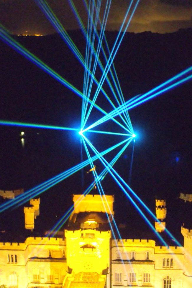 Bright blue and green laser beams radiate from a dark sky, enhanced by an incredible drone show, behind an illuminated castle at night.