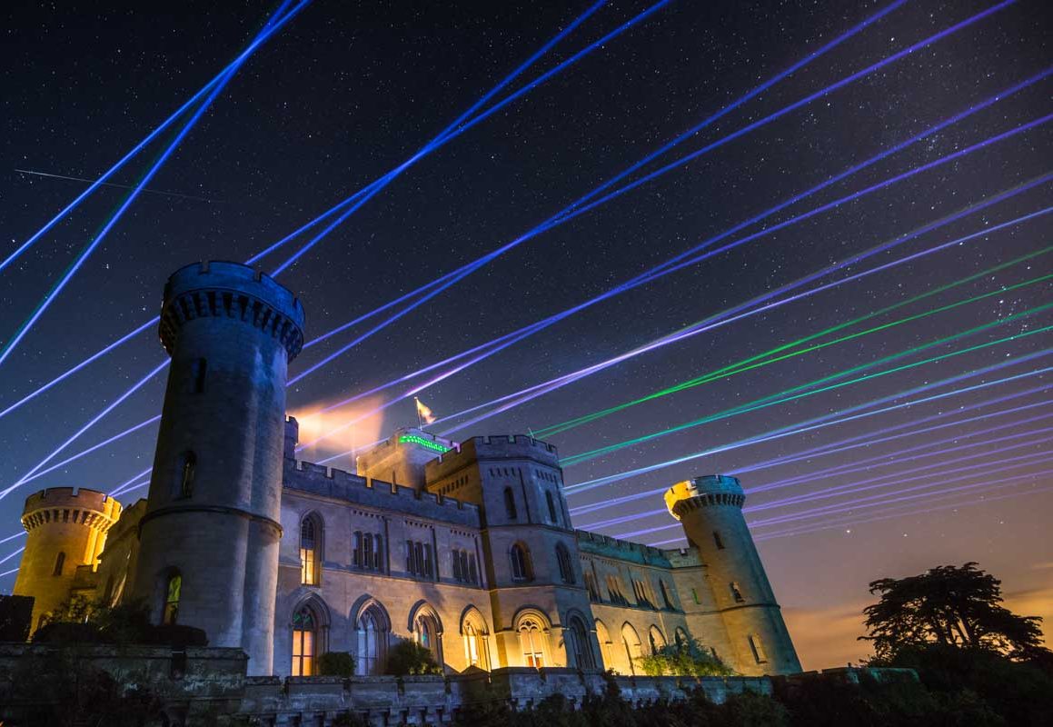A historic castle illuminated at night with colorful laser beams crossing the sky, enhanced by a mesmerizing drone show, with stars visible above.