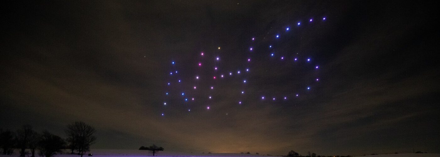 A mesmerizing drone light show illuminates the night sky as drones form "NHS" in colorful lights, against a landscape adorned with silhouetted trees.