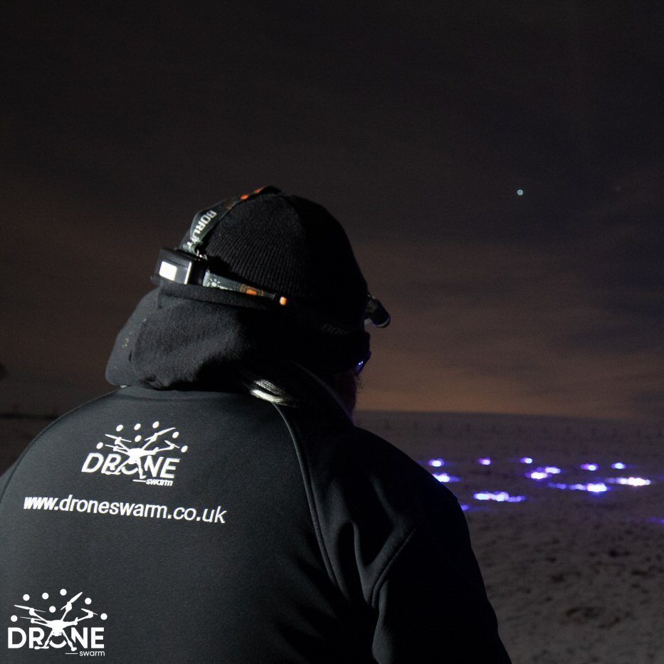 Person in warm clothing with "droneswarm.co.uk" on their back overseeing a mesmerizing drone light show, with blue lights illuminating the night sky.