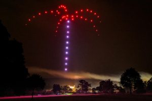 A mesmerizing drone show creates a firework-like pattern in the night sky above a lit building, with trees in the foreground adding to the enchanting scene.