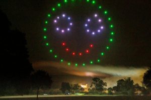 A glowing smiley face made of drones illuminates the night sky, transforming the dark landscape with trees and distant lights into a mesmerizing drone show.