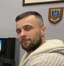 A man with short hair and a beard wearing a light-colored sweater smiles slightly. A plaque is visible in the background, hinting at his recent success at organizing an impressive drone show.