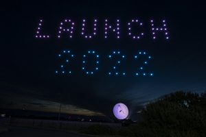 A mesmerizing drone show lights up the night sky as drones form "LAUNCH 2022," with a large balloon glowing on the ground below.
