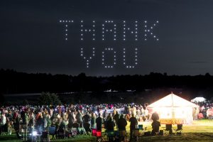 A large crowd gathers at night for a mesmerizing drone show, watching in awe as drones form the words "THANK YOU" against the dark sky.