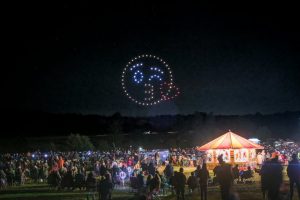 A crowd watches a mesmerizing drone show at night, with an animated smiley face and heart brilliantly formed by drones in the sky.