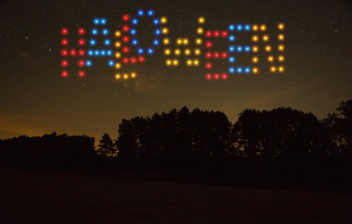 Lights in the night sky form "MOBILE!" in red, blue, and yellow, above a dark silhouette of a forest during a mesmerizing drone light show.