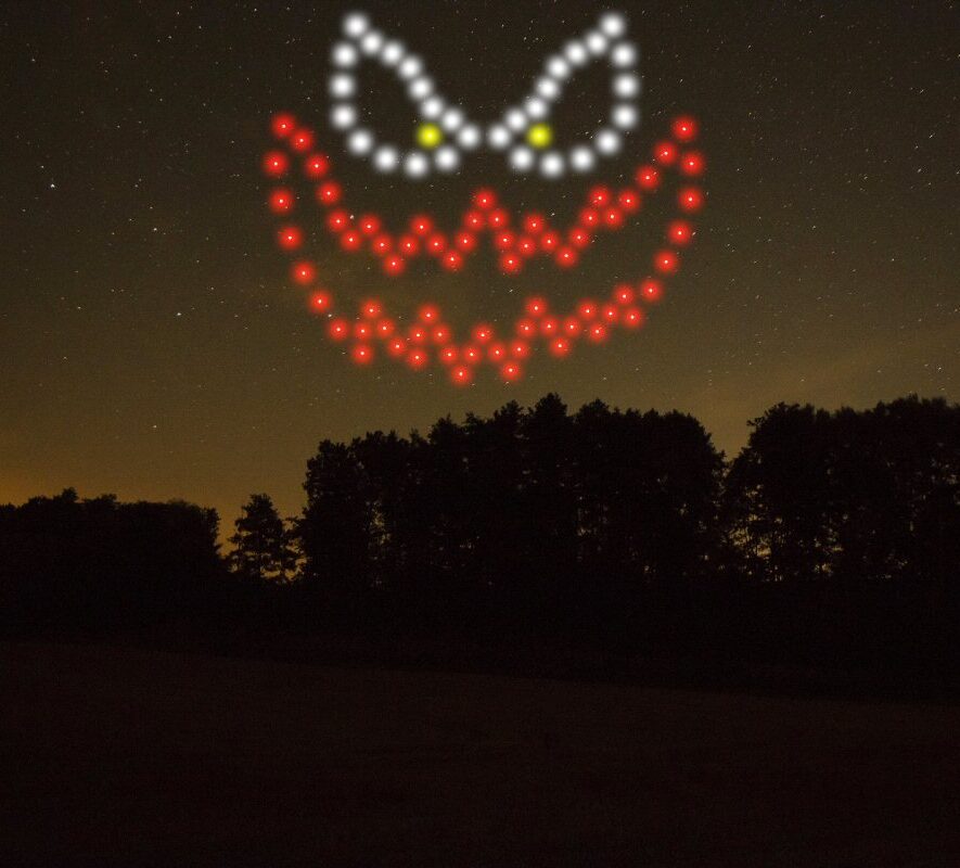 A night sky displays a spooky pumpkin face made of red, white, and yellow lights above a line of silhouetted trees, part of an enchanting drone light show.