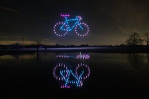 A drone show lights up the night sky, drones forming a glowing bicycle shape that reflects in the still lake below.