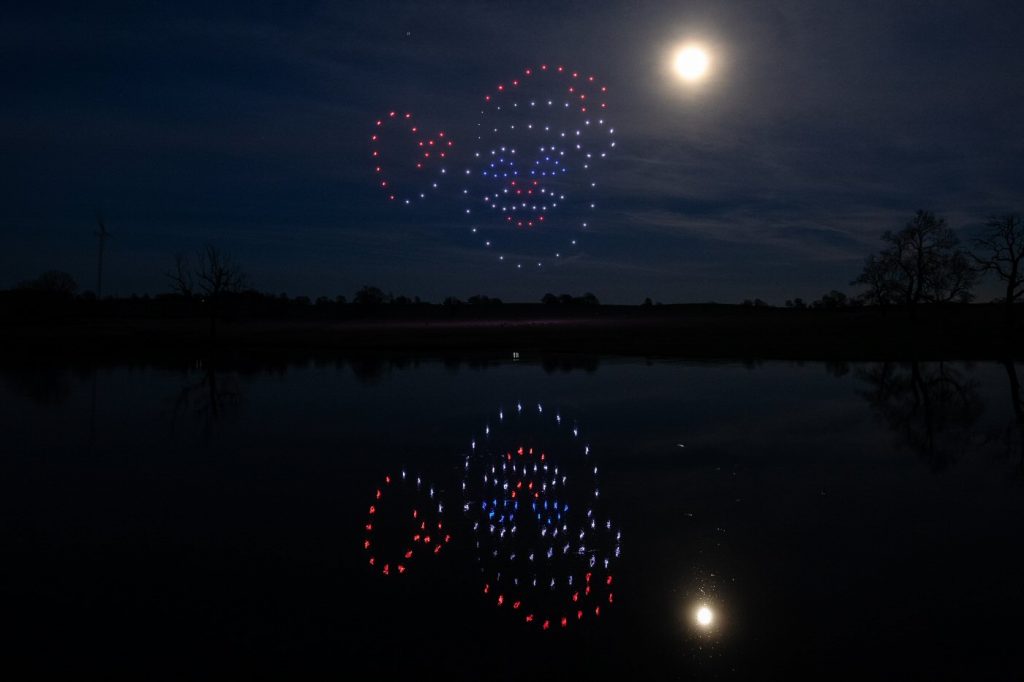 A night sky showcases a mesmerizing drone light show of a smiling face with a hat, its vibrant reflection shimmering in the calm lake below.