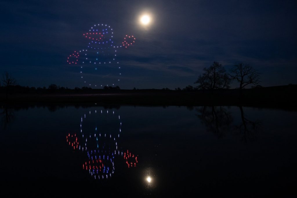 A drone light show forms a snowman in the night sky, its shimmering reflection dancing on a calm lake under a full moon.