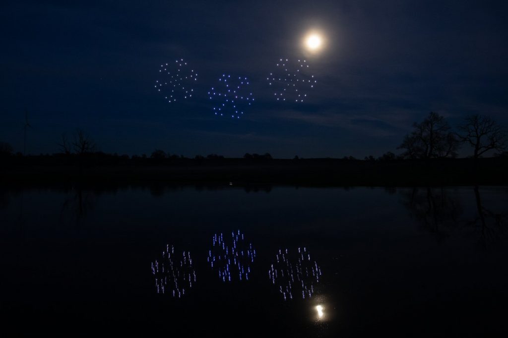 Drones form patterns resembling fireworks in the night sky, creating a stunning drone light show with the moon shining brightly and reflections on a lake.
