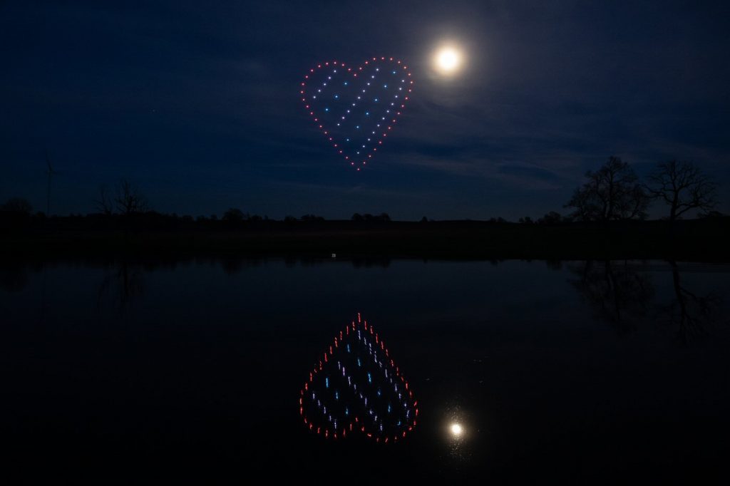A nighttime scene with a moon and a heart-shaped light display, created by an enchanting drone light show in the sky, reflected in a calm body of water.