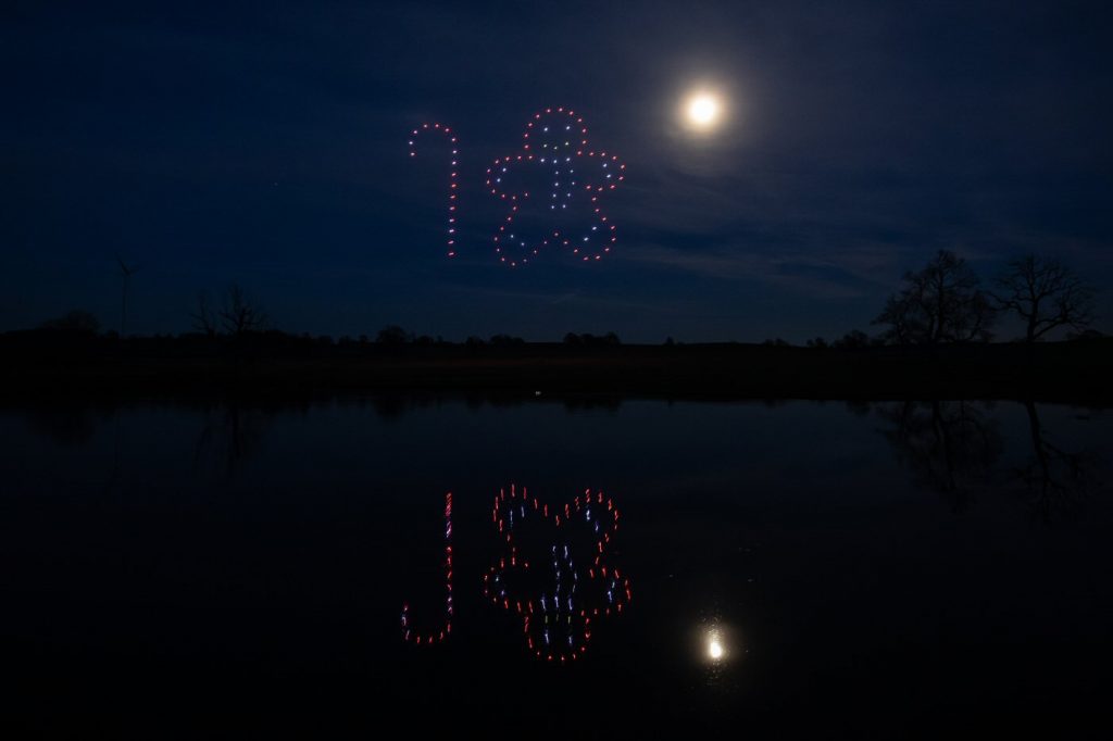 Drones form a candy cane and gingerbread man shape in the night sky above a lake with a full moon, creating an enchanting drone light show.