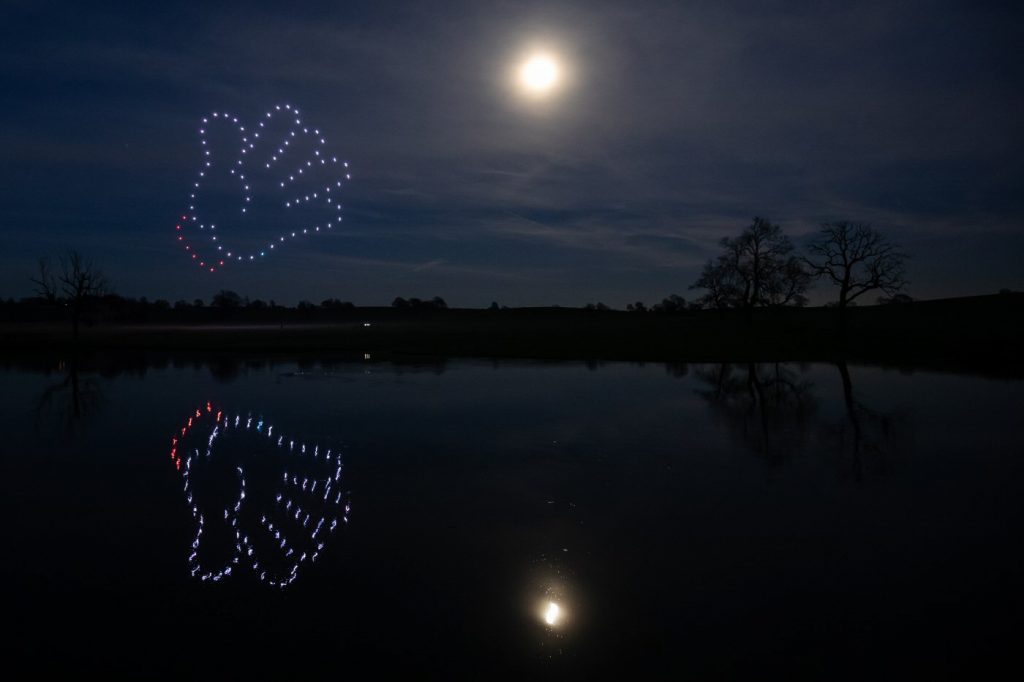 A nighttime scene with a full moon; a mesmerizing drone light show forms two hands, mirrored over a calm lake.
