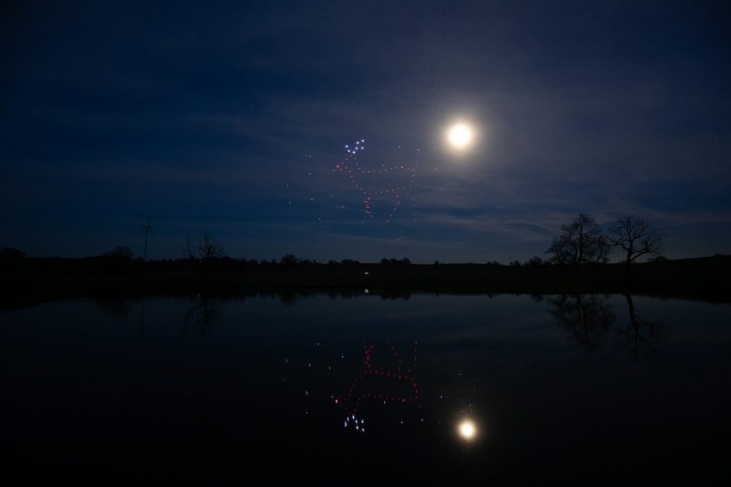 Night sky with a full moon, a drone light show forming a reindeer pattern, and a reflection on the calm lake below.