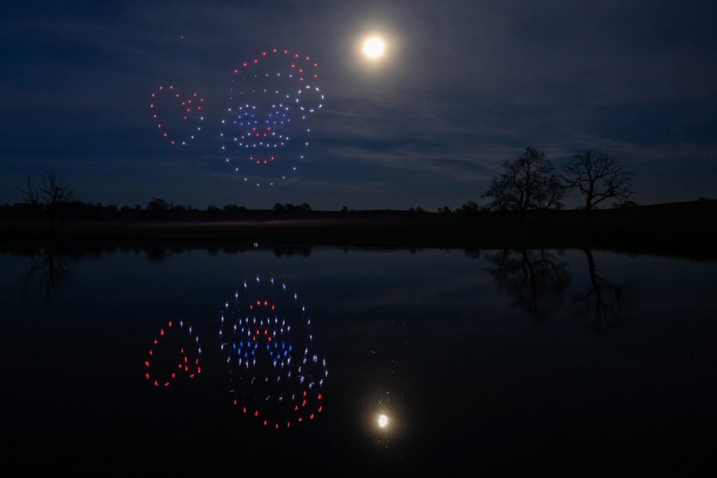 A night sky with a full moon and a mesmerizing drone light show forming heart and face-shaped patterns, all beautifully reflected in the calm lake.