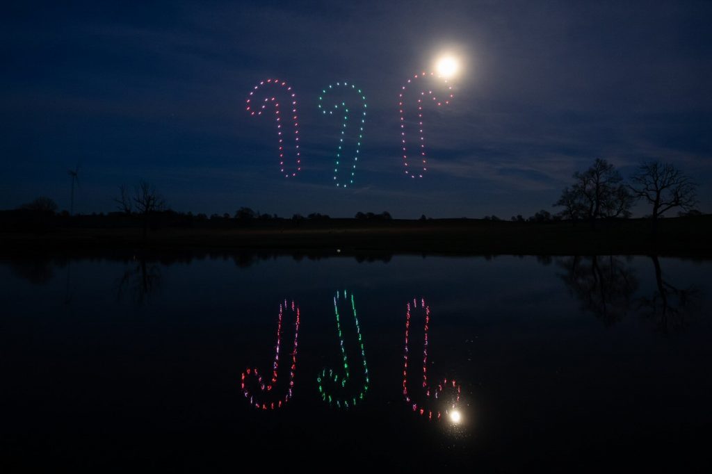 Candy cane-shaped lights, part of a drone light show, illuminate the night sky above a calm lake, reflecting the shapes in the water below.