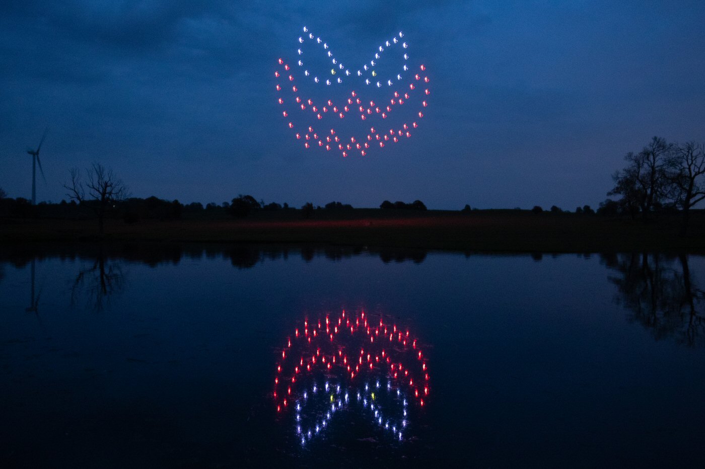 A mesmerizing drone light show depicts a glowing jack-o'-lantern face in the night sky, perfectly reflected in the calm body of water below.