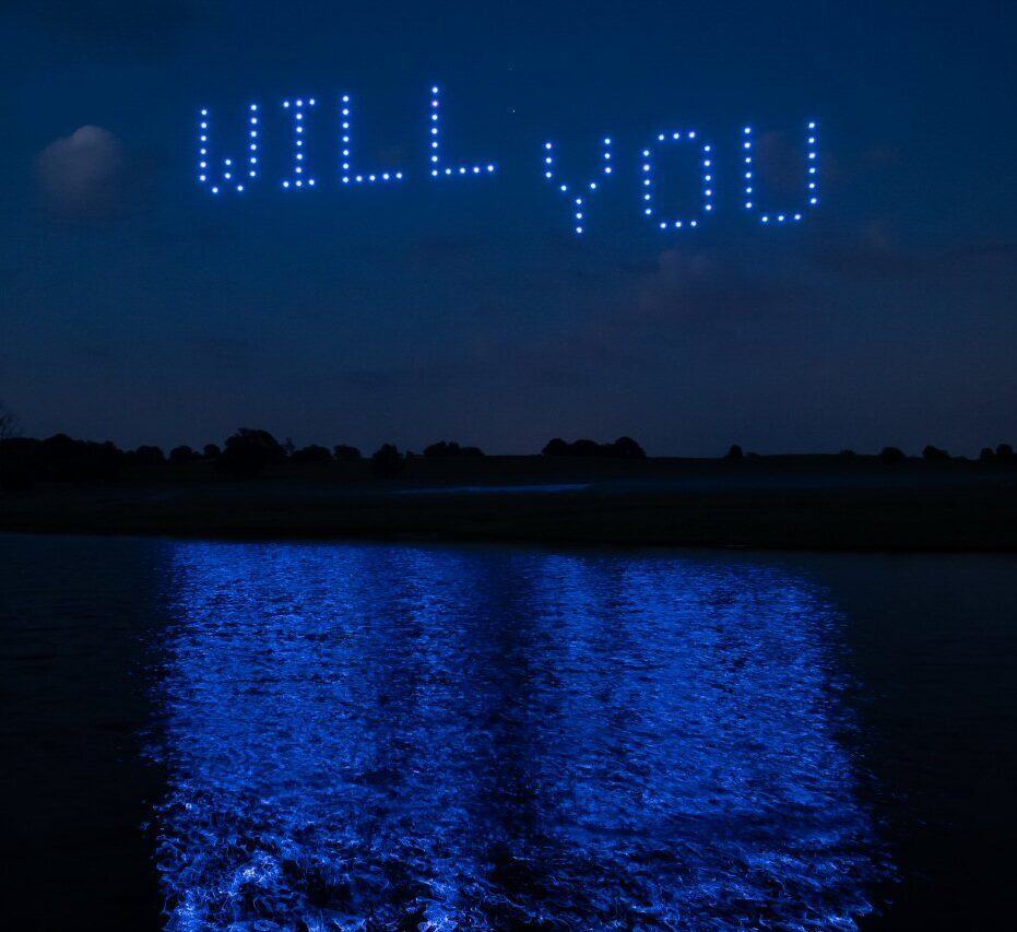 A night sky features a captivating drone light show with the words "WILL YOU" formed by drones, perfectly reflected on the water surface below.