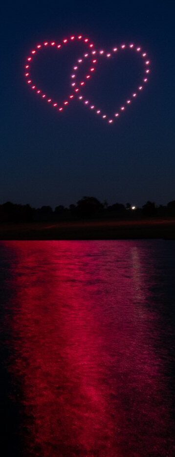 Two illuminated heart shapes in the night sky, created by a stunning drone light show, reflecting on a body of water below.