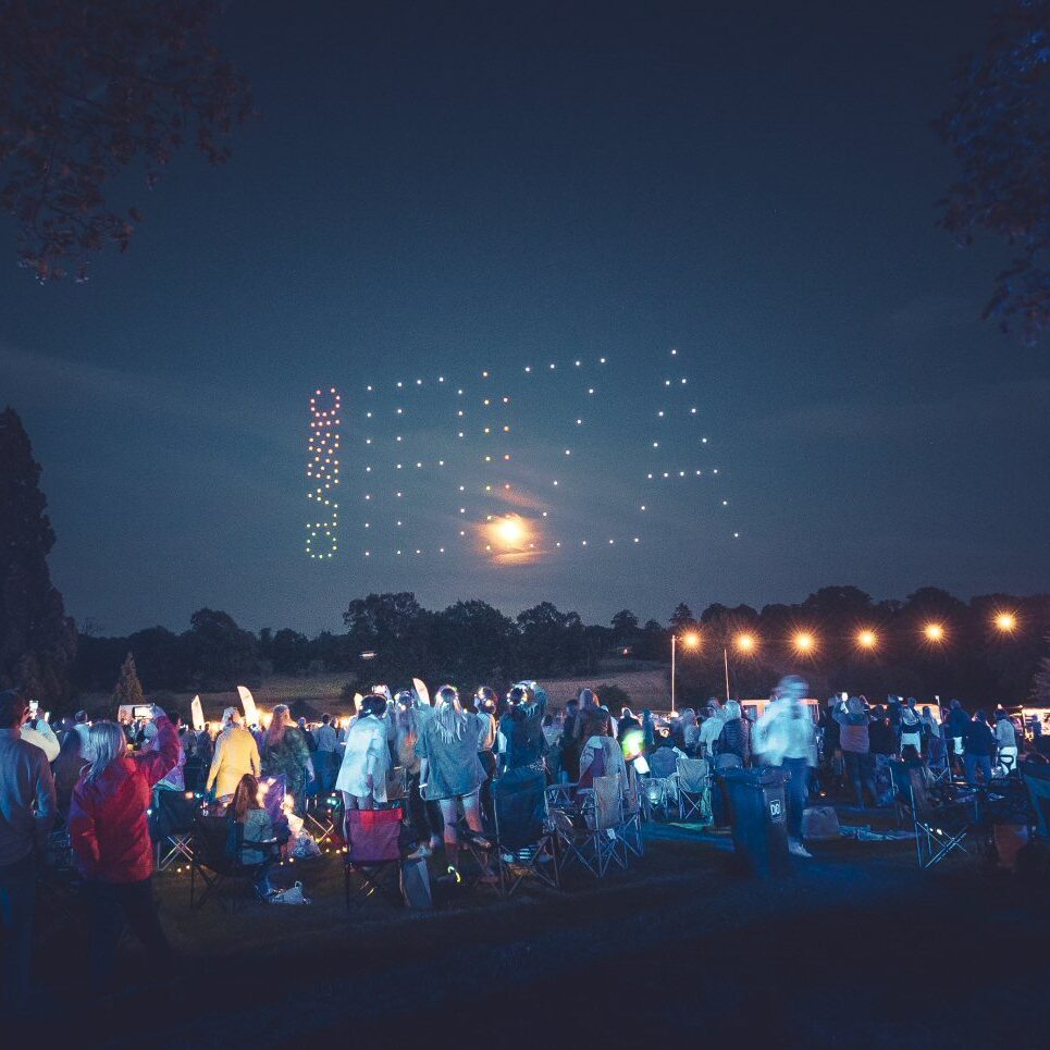 Crowd at an outdoor event at night, mesmerized by a captivating drone light show forming "IBIZA" in the sky.