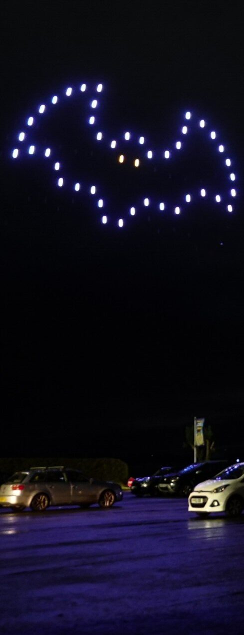 A stunning drone light show forms a bat symbol in the night sky, with cars parked below in awe.