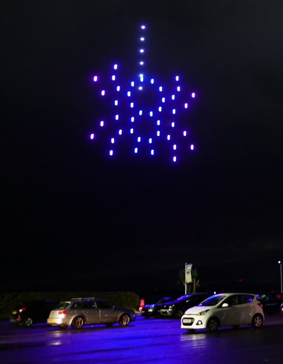 A mesmerizing formation of blue and purple lights danced in the night sky above a car park, casting an enchanting glow over the parked cars, like a drone light show painting the heavens.