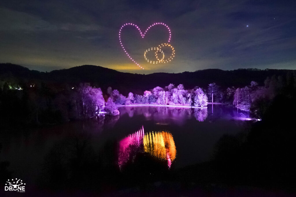 A mesmerizing drone light show shapes a heart and two rings above a forested lake, their vibrant pink and yellow lights reflecting brilliantly on the water.