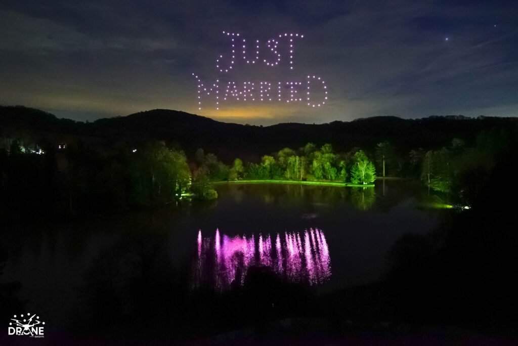 A drone light show creates a "Just Married" display in the night sky over a lake surrounded by illuminated trees and hills.