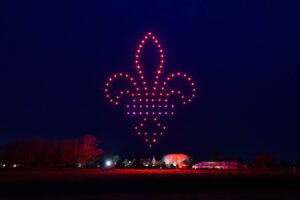 A mesmerizing drone light show displays a glowing red fleur-de-lis in the night sky above a rural landscape with houses and trees.