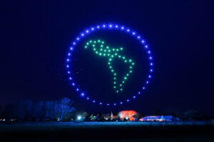 A mesmerizing drone light show creates a circle of blue and green lights in the night sky, forming a globe map of the Americas over a dark landscape.