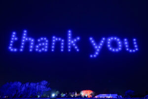 A night sky displays the words "thank you" formed by blue lights, courtesy of an enchanting drone light show, above a lit-up landscape.
