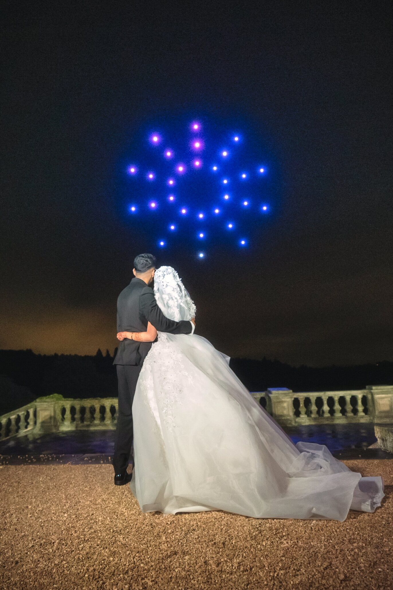 A bride and groom with backs turned, watching a light show in the night sky at an outdoor venue with a stone railing.