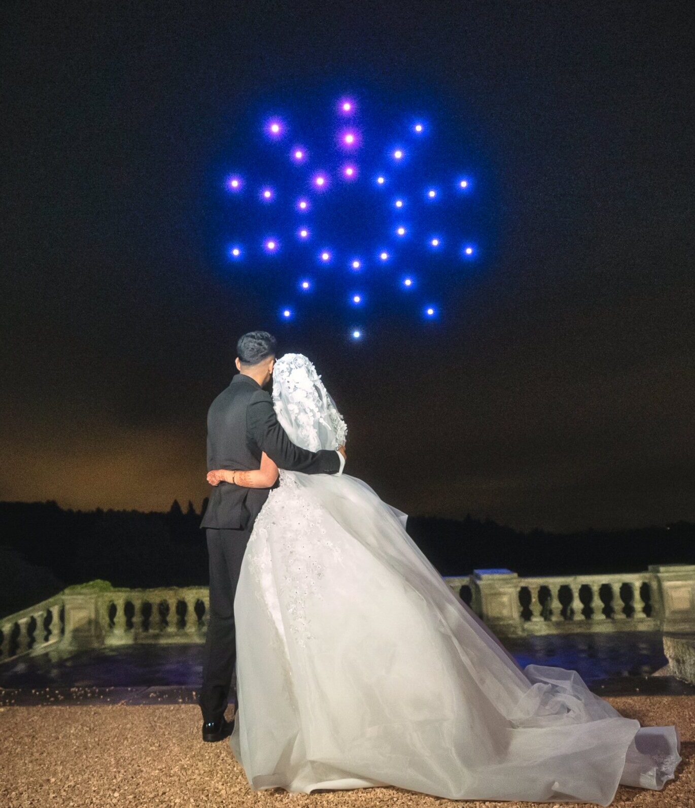 A bride and groom with backs turned, watching a light show in the night sky at an outdoor venue with a stone railing.