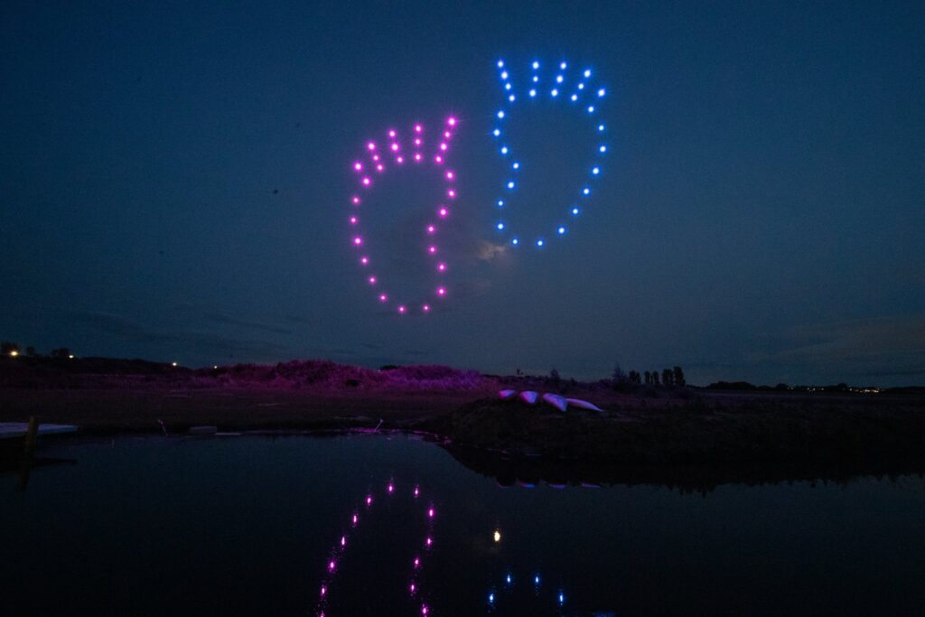 Gender Reveal Drone Light Show Display - footprints made of pink and blue drone lights are displayed in the night sky above a calm landscape.