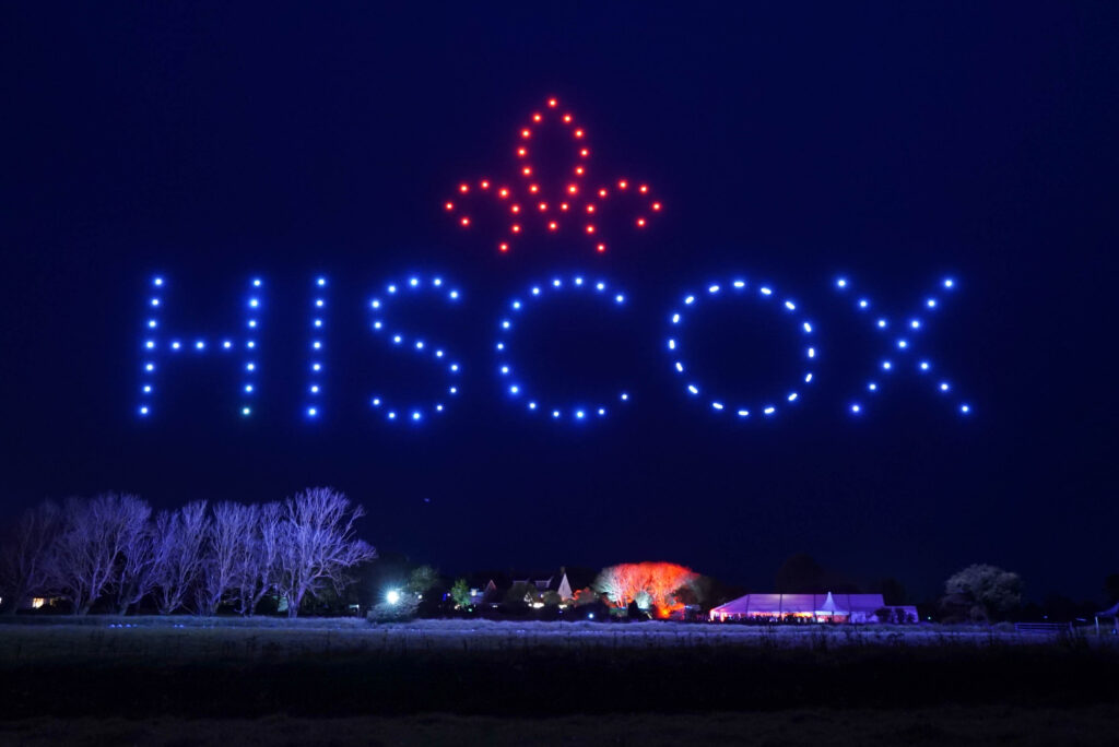 Drones form the word "HISCOX" and a red fleur-de-lis in the night sky above trees and buildings.