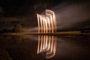 Light trails soar above a grassy field at night, reflected in a calm pond under a dark sky.