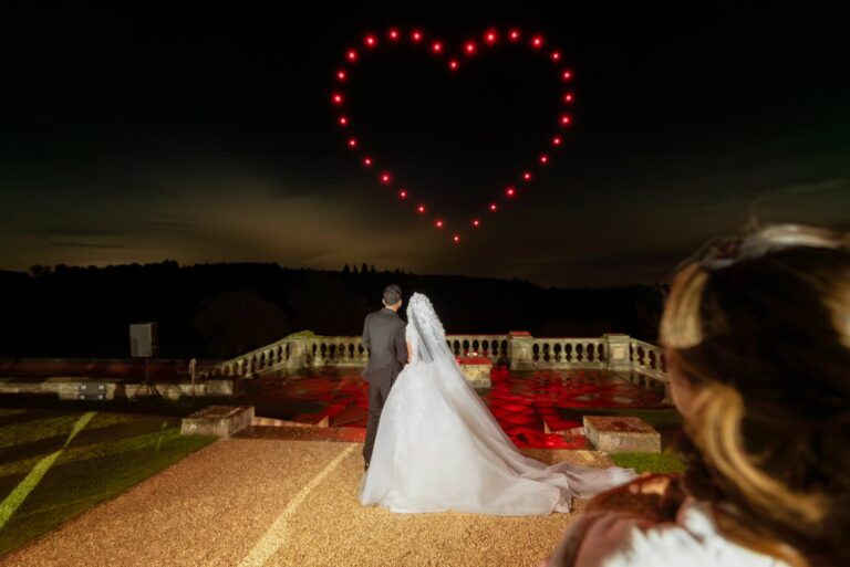 A bride and groom watch heart-shaped red drone lights in the night sky at their outdoor wedding.