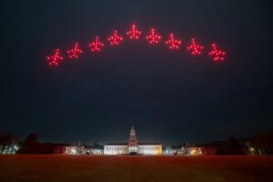 Red drones form airplane shapes in an arc above a lit building at night.