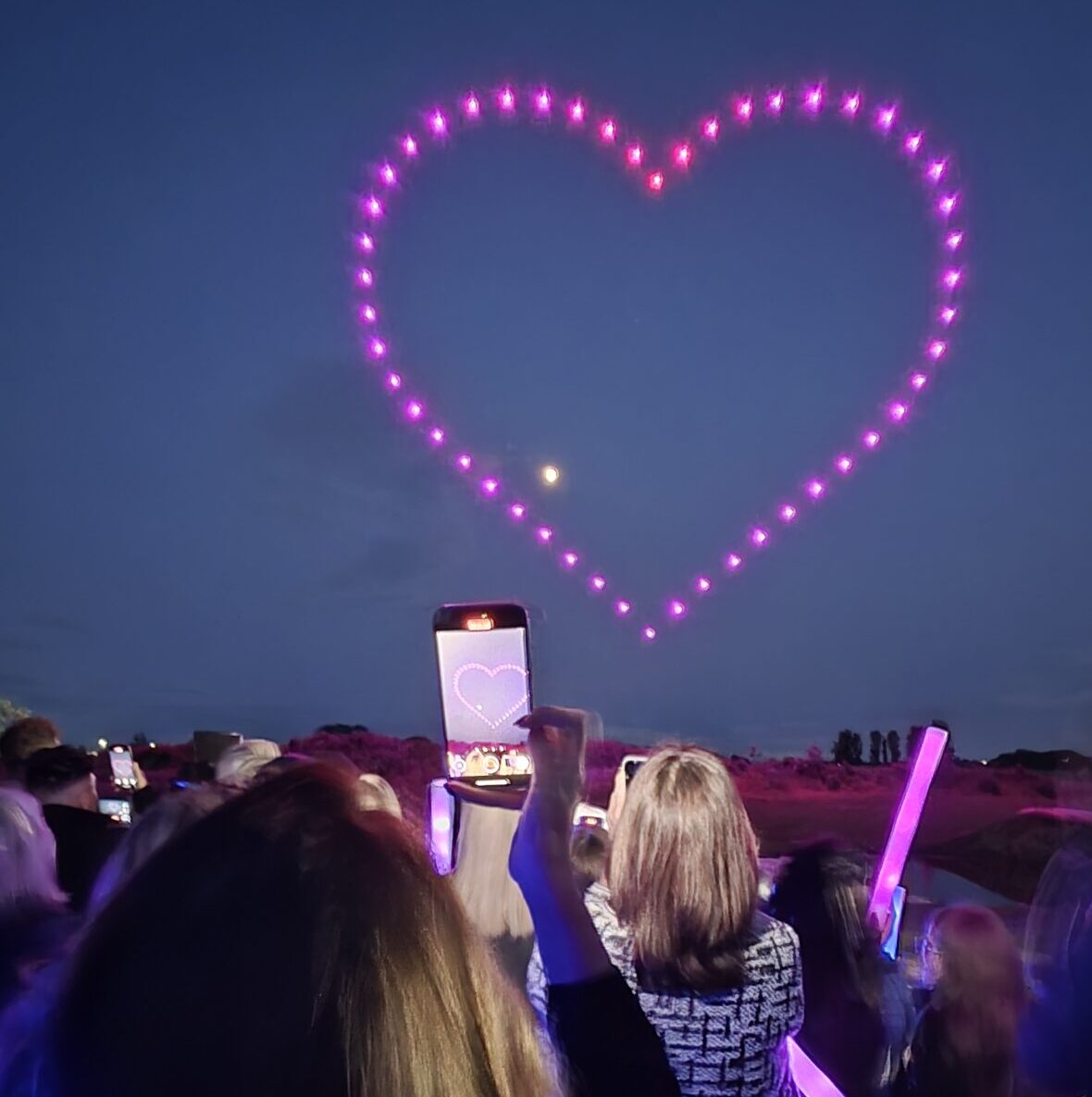 A glowing pink heart shape made by synchronized drones lights up the night sky, while crowds gather below, watching in awe and capturing the magical moment on their phones.