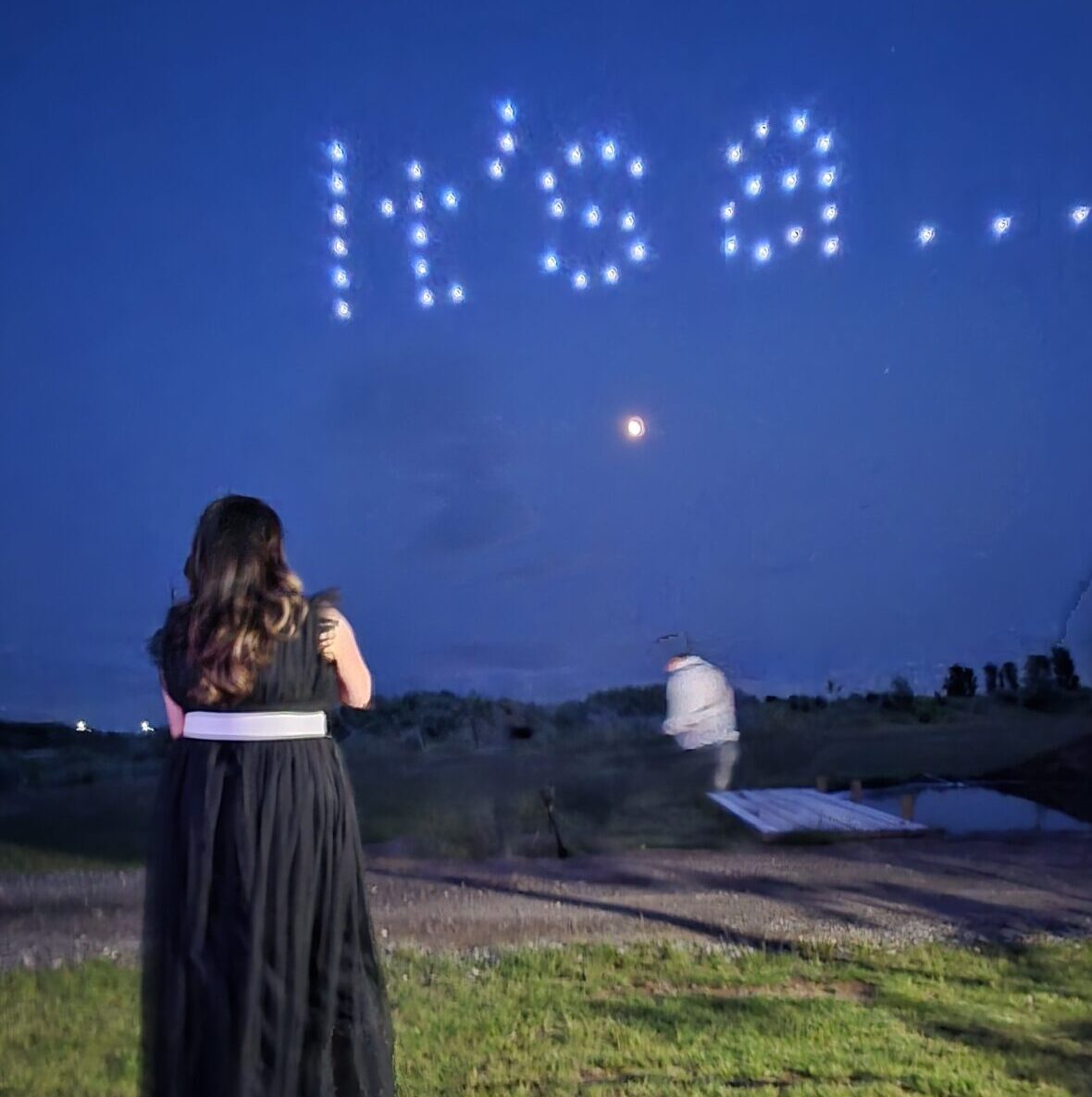 Woman in a black dress gazes upward as drones illuminate the night sky, spelling out "It's a..." during an unforgettable gender reveal.