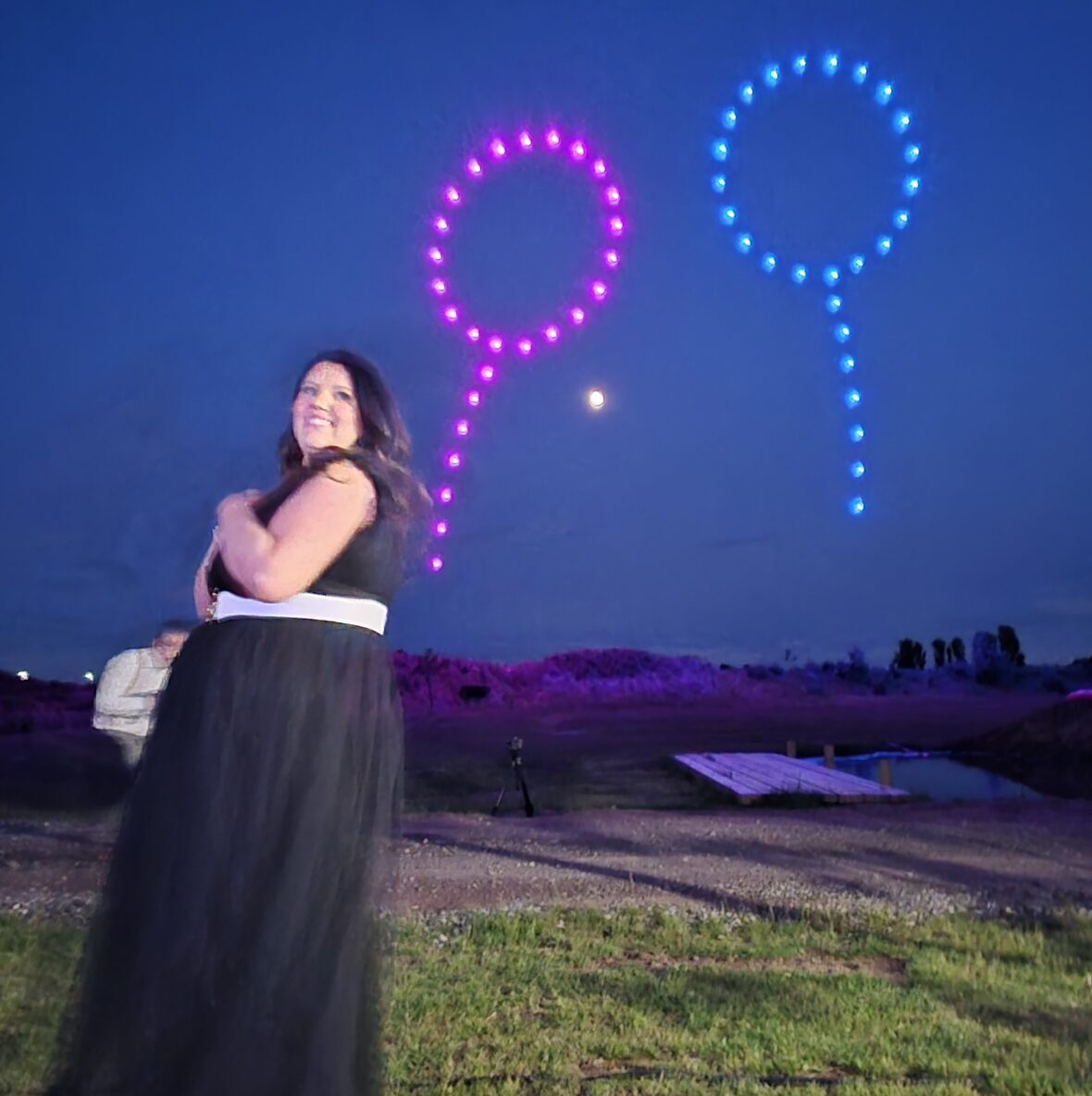 A woman in a black dress stands outside at night, mesmerized as colorful drones dance above, forming pink and blue shapes in the starry sky.