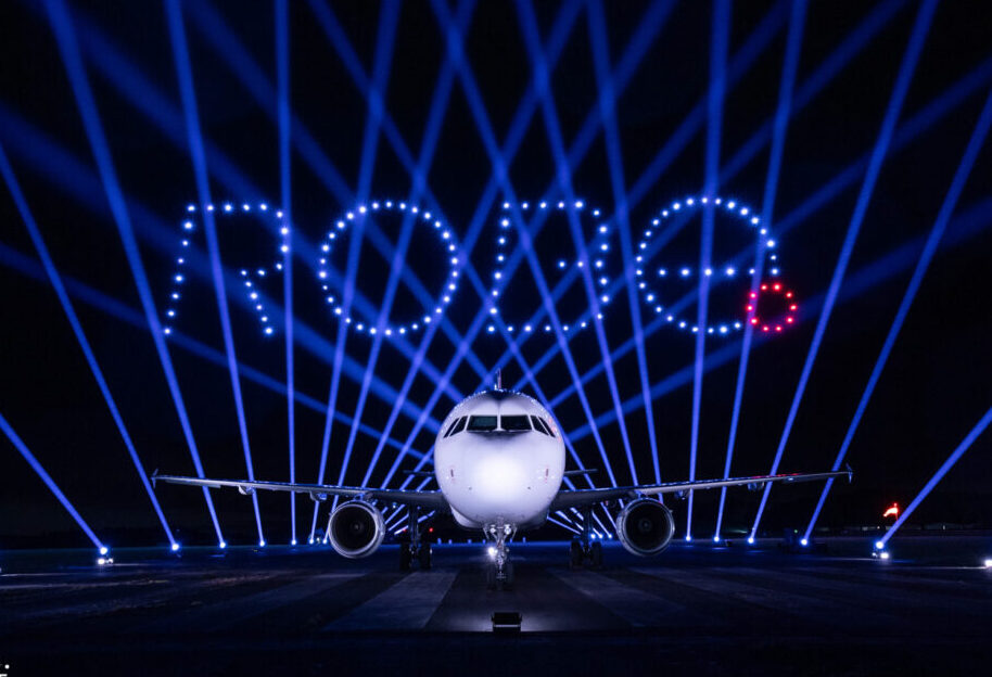 A plane on a runway at night with "ROUGE" spelled in lights, beams shining in the background, and a dazzling DroneSwarm illuminating the scene.
