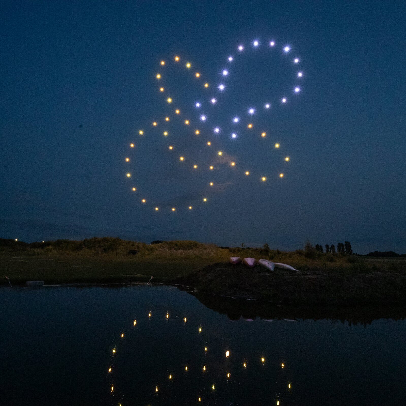 Drones form glowing shapes in the night sky for a spectacular gender reveal, their lights reflecting on a calm body of water below.