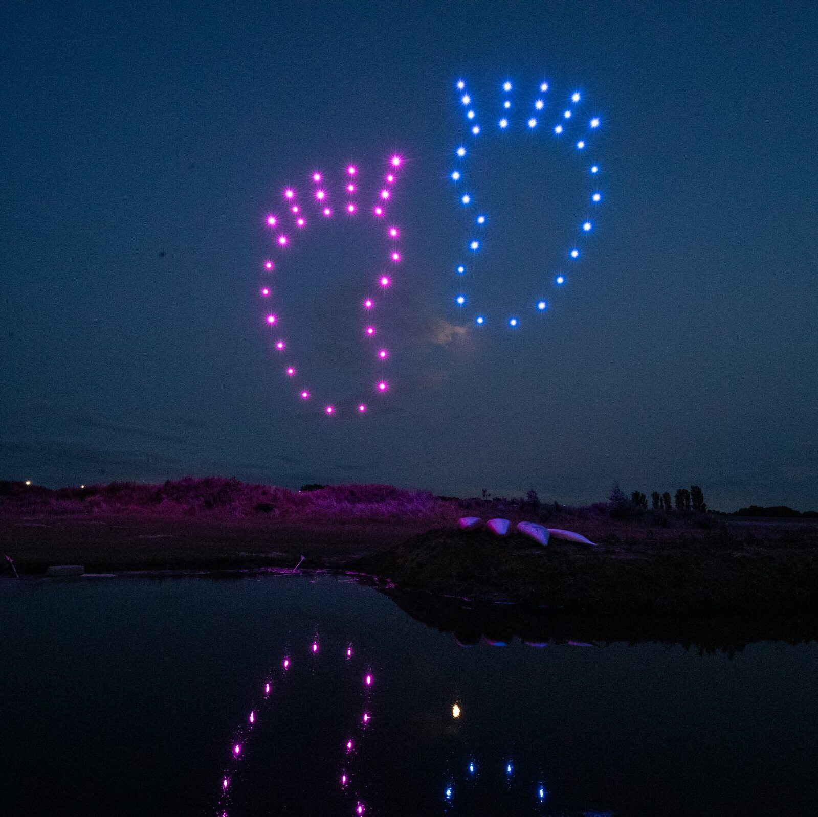 Pink and blue drones form the shapes of baby feet in the night sky, reflected in calm water below—a stunning gender reveal display.