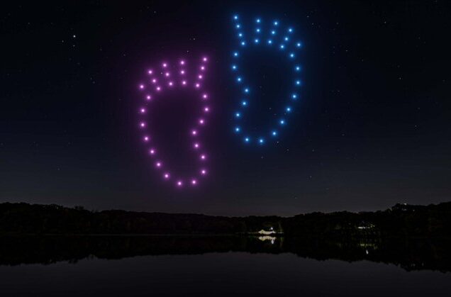 A vibrant droneswarm paints purple and blue baby footprints in the night sky above a shadowy landscape.