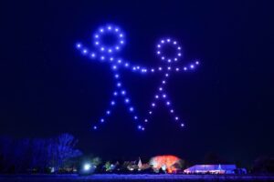 Blue drones form stick figures holding hands in the night sky above trees and buildings.