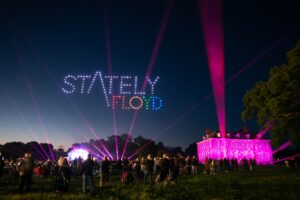 A crowd watches a drone light display spelling "Stately Floyd" at night, with colorful lights on a mansion.